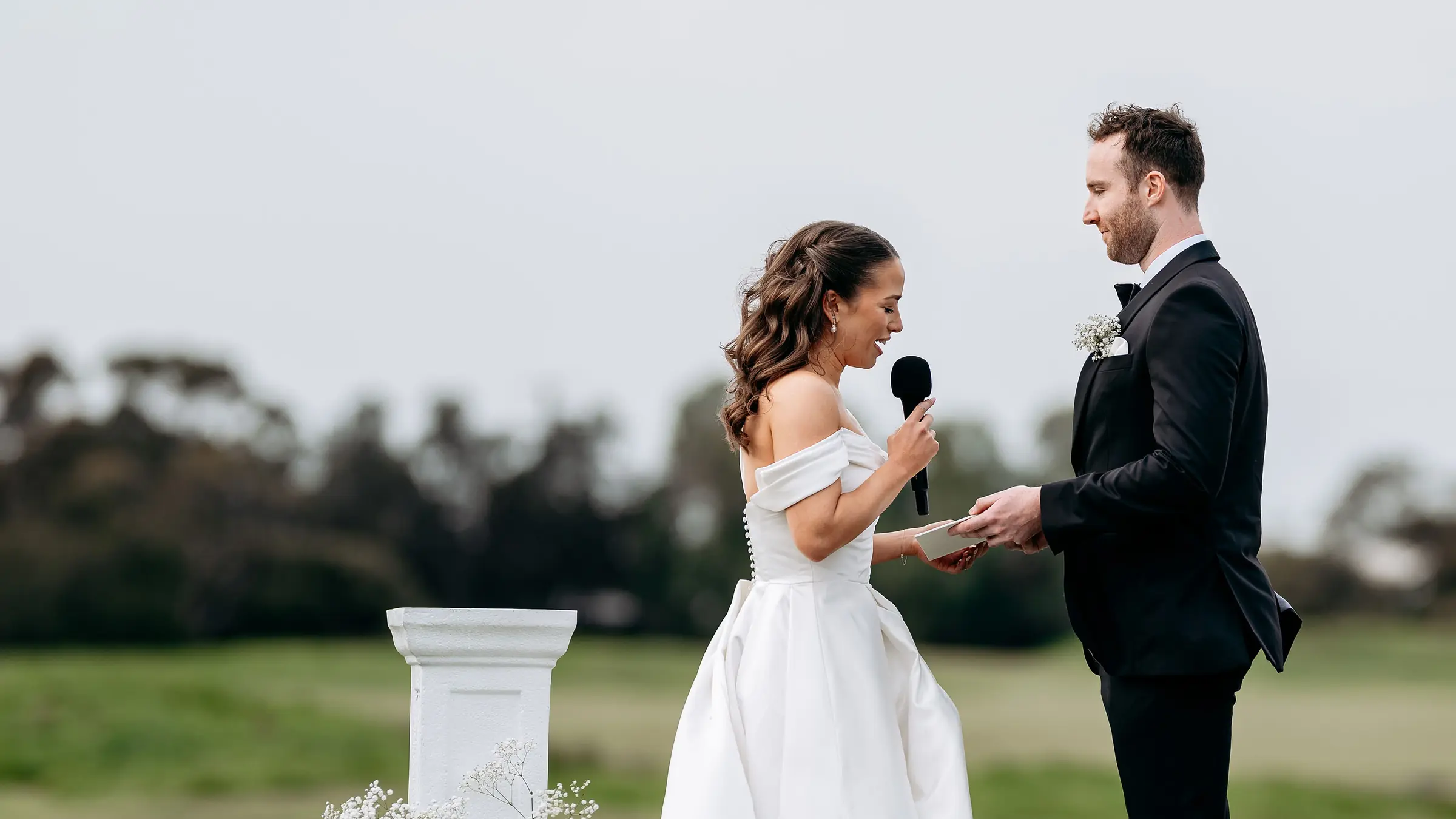 A bride reading her vows into a microphone to her groom on a fairway, between urns of baby's breath