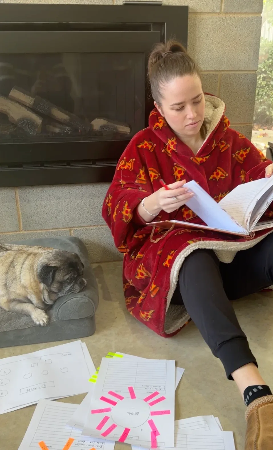 Elise sitting on the floor by a fireplace in a red hooded blanket, reading through a planning book with a pen in hand. A pug sleeps on a dog bed alongside, and printed seating plans cover the floor in front: a hand-drawn room layout and round-table plans with a sticky note for each seat.